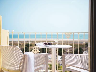 Balkon mit weißen Stühlen und Tisch, Meer im Hintergrund unter blauem Himmel.
