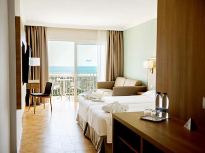 Hell eingerichtetes Hotelzimmer mit Doppelbett, Sofa, Schreibtisch und Meerblick vom Balkon.