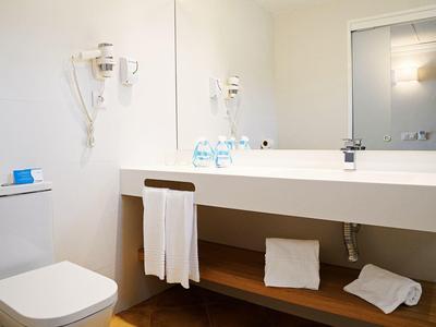 Helles Badezimmer mit weißem Waschbecken, Handtüchern, Toilettenpapier und einem Spiegel an der Wand.