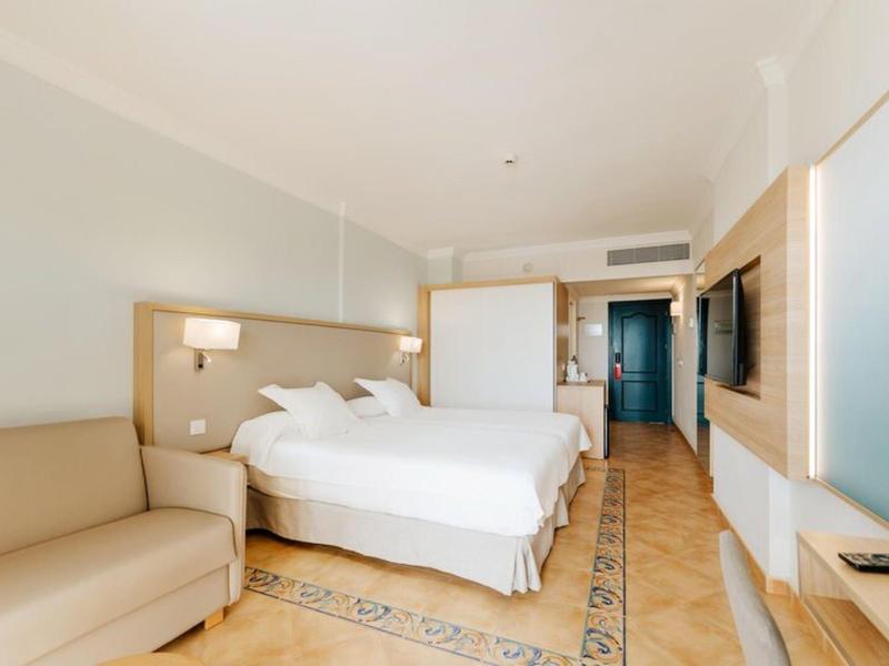 Camera d'albergo luminosa con due letti singoli, un divano beige, pavimento piastrellato decorato e TV a parete.