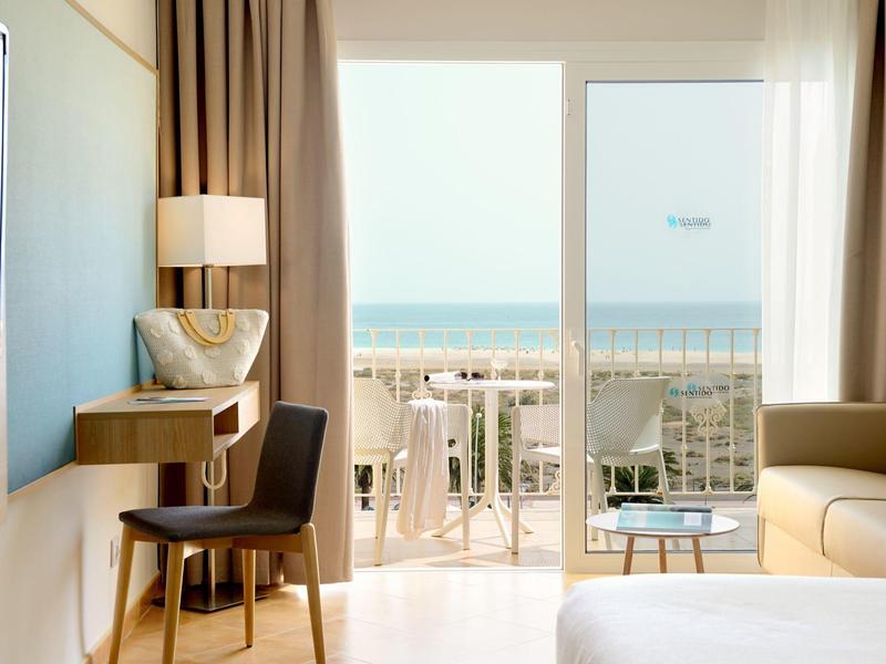 Camera d'hotel luminosa con vista mare, scrivania, sedia, lampada, divano e balcone con sedie esterne.