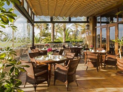 Zastřešená terasa s dřevěnými stoly a židlemi, obklopená rostlinami a palmami.