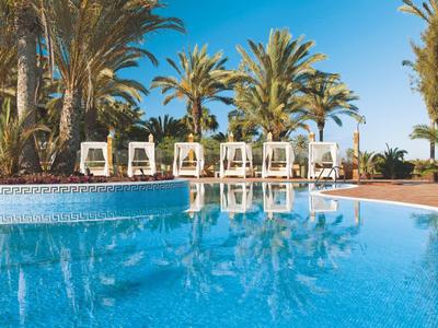 Blauer Pool mit weißem Rand, Sonnenliegen und Palmen unter blauem Himmel.