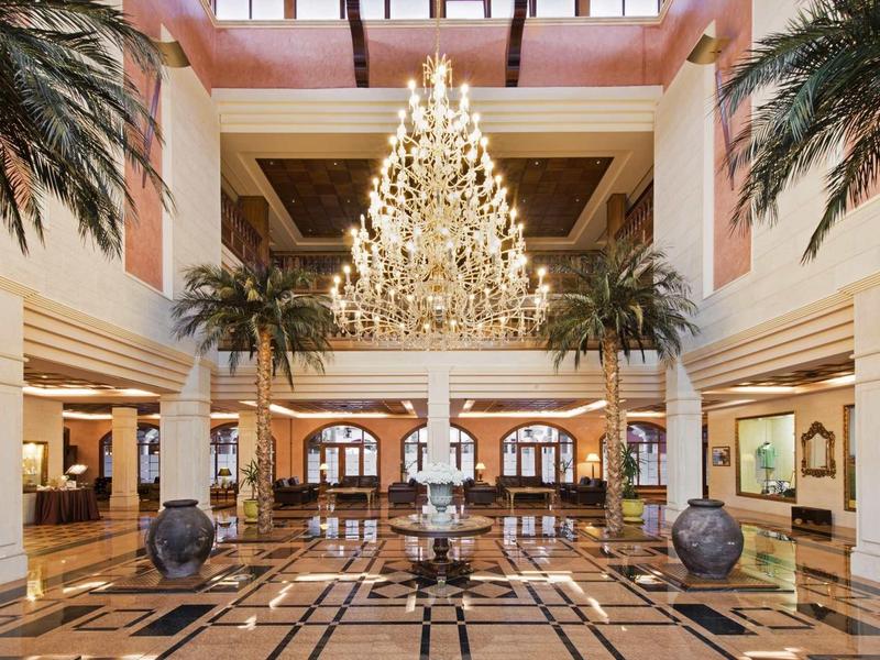 Velká hotelová lobby s lustrem, mramorovou podlahou a dekorativními palmami.