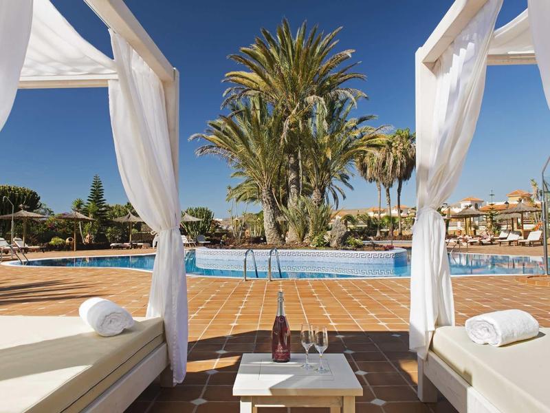 Výhled z bílé bazénové lounge se závěsy na bazén a palmy pod modrou oblohou.