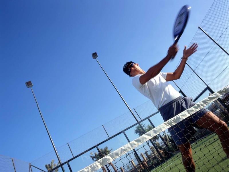 Muž hrající paddle tennis na slunném kurtu s jasnou modrou oblohou.