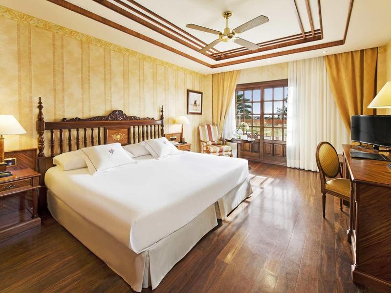 Elegantní hotelový pokoj s velkou postelí, dřevěnou podlahou a okny s výhledem na terasu.