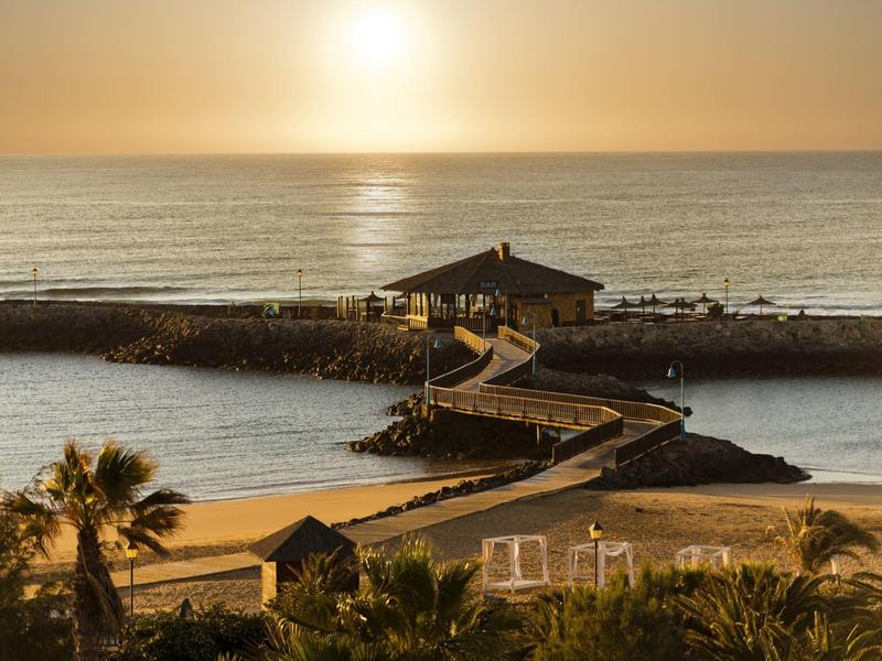 Un camino conduce a un pequeño edificio junto al mar al atardecer con palmeras en primer plano.
