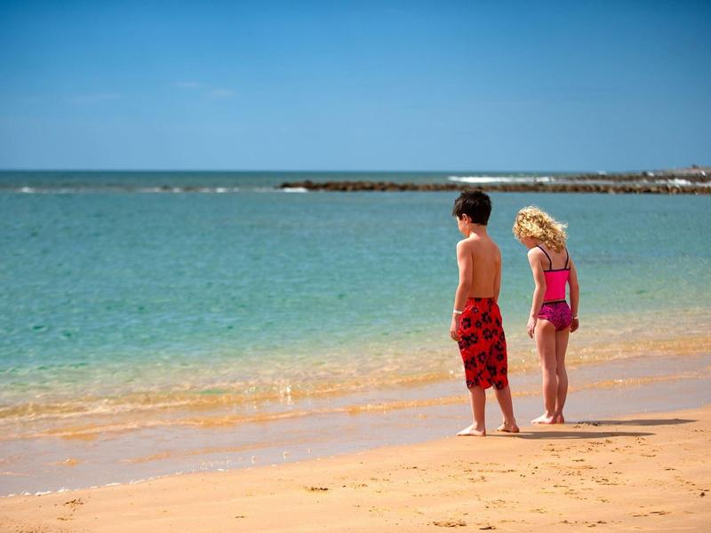 Dos niños están en la playa de arena mirando al mar tranquilo bajo un cielo despejado.