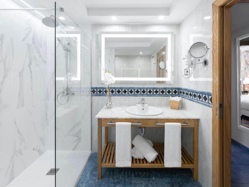 Baño luminoso con ducha, espejo iluminado y mueble bajo de madera con toallas.