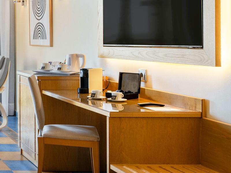 Habitación de hotel luminosa con mesa de madera, silla, televisor y cafetera.