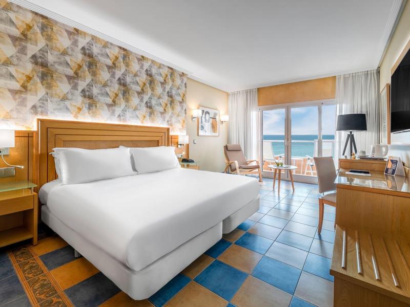 Habitación de hotel luminosa con grandes ventanas, vista al mar, cama doble e interior moderno.