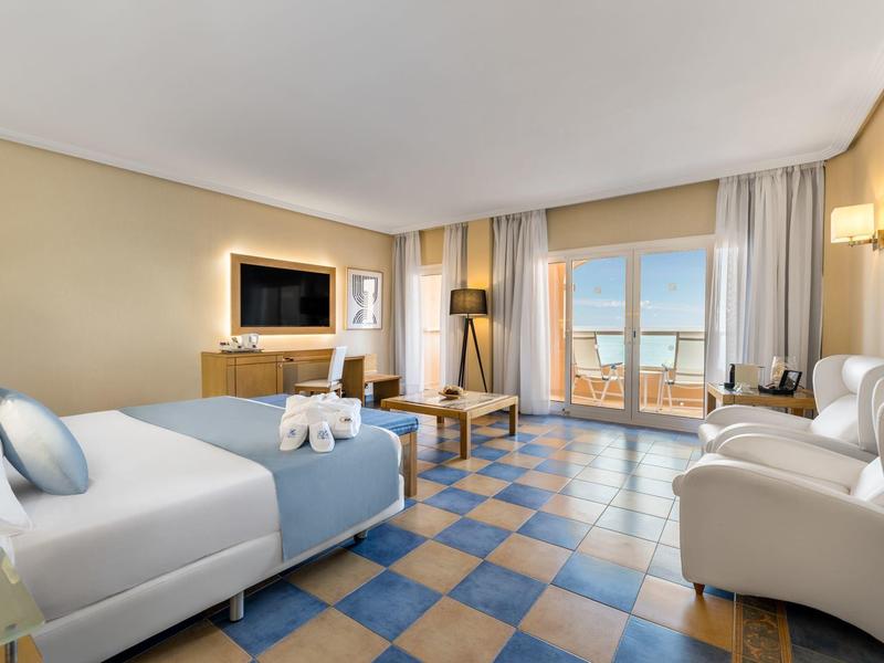 Sala luminosa con azulejos azules y beige, sofá blanco y vista al mar a través de grandes ventanas.