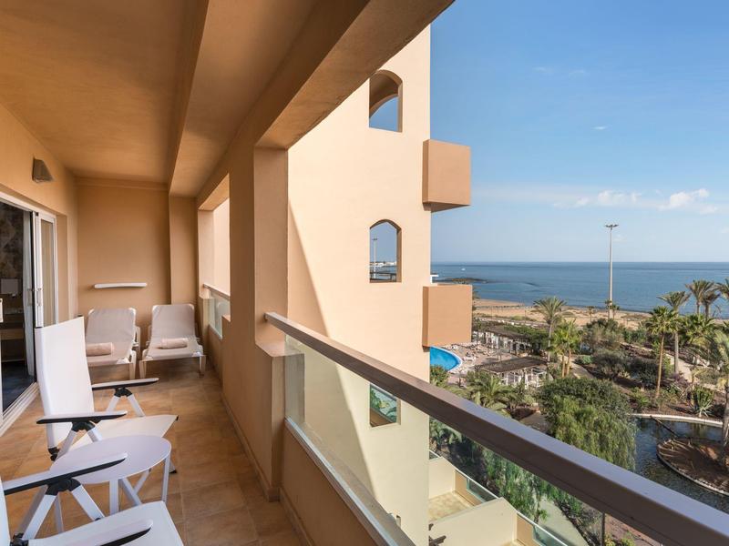 Balcón con sillas blancas y vista a jardín, piscina y mar bajo un cielo despejado.