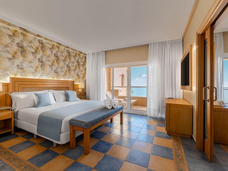 Habitación de hotel luminosa con cama doble, muebles de madera, balcón y vista al mar en clima soleado.