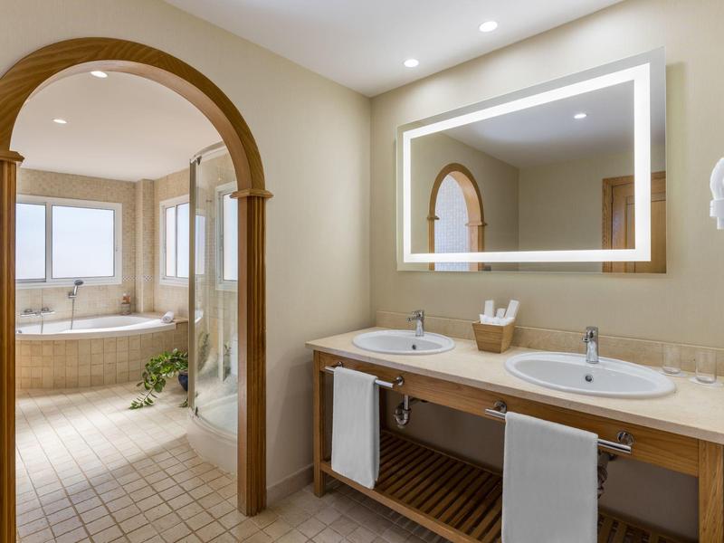 Baño elegante con lavabo doble, gran espejo iluminado y bañera al fondo.