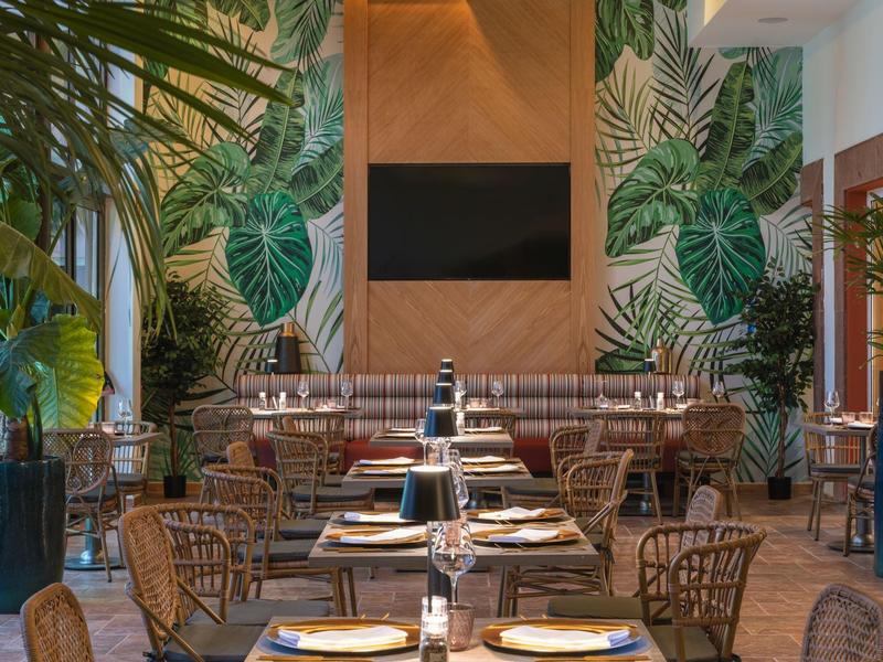 Restaurante elegante con pared de hojas, mesas y sillas de madera bajo luz cálida.