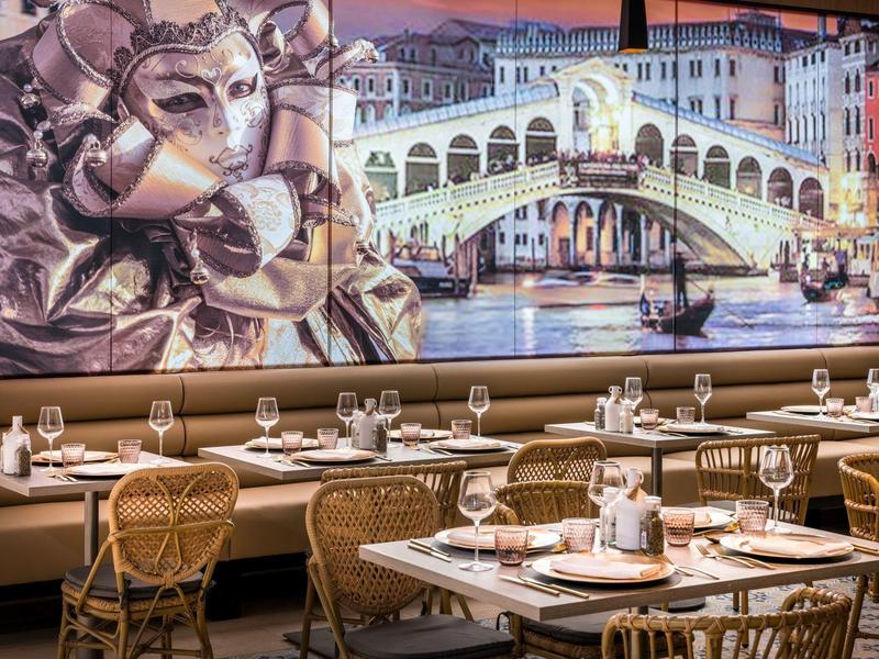 Restaurante elegante con mesas puestas, sillas de mimbre y gran mural de puente veneciano