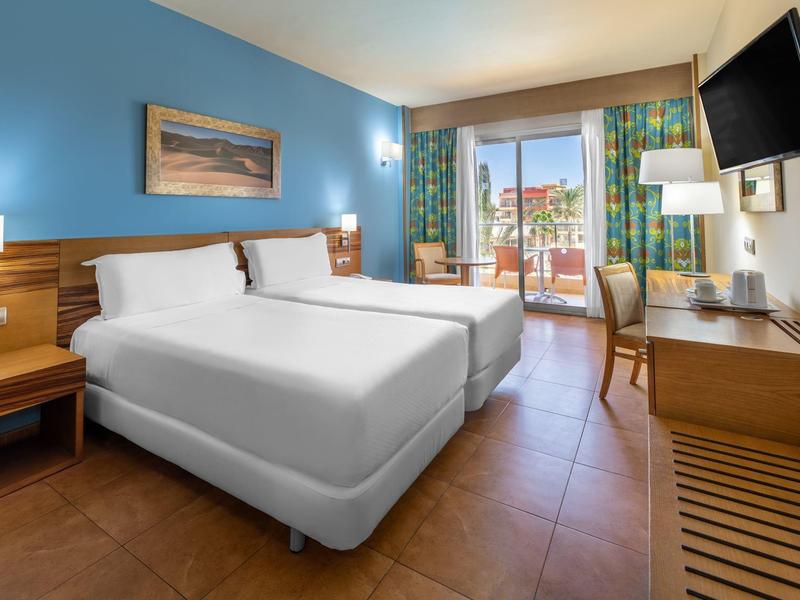 Lichte hotelkamer met twee bedden, houten meubels en balkon met zeezicht.