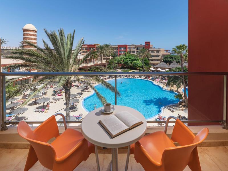 Balkon met twee oranje stoelen en een klein tafeltje met uitzicht op zwembad en hotel bij zonnig weer.