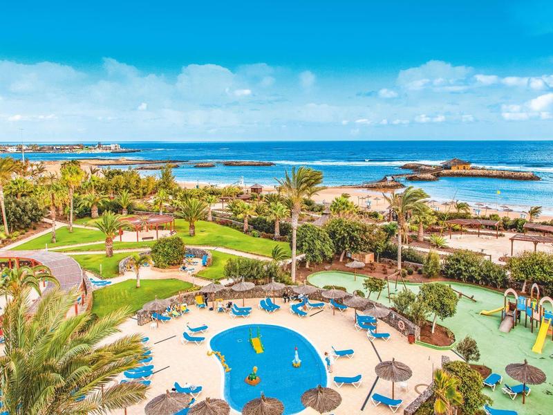 Hotel tuin met zwembad, parasols, palmbomen en zeezicht onder een blauwe hemel.