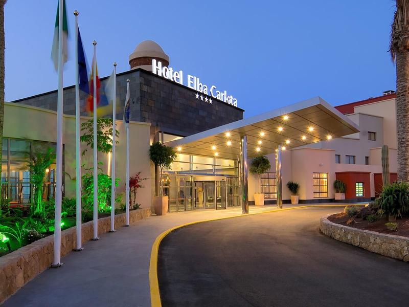 Verlichte hotelentree met vlaggen en palmbomen bij schemering.