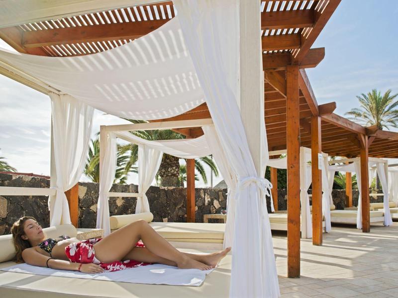 Vrouw ontspant op een ligstoel onder een houten pergola met witte gordijnen.