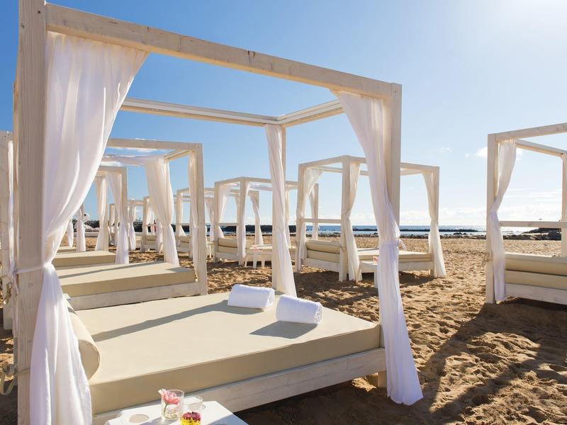 Witte ligbedden met gordijnen op zandstrand onder een heldere lucht.