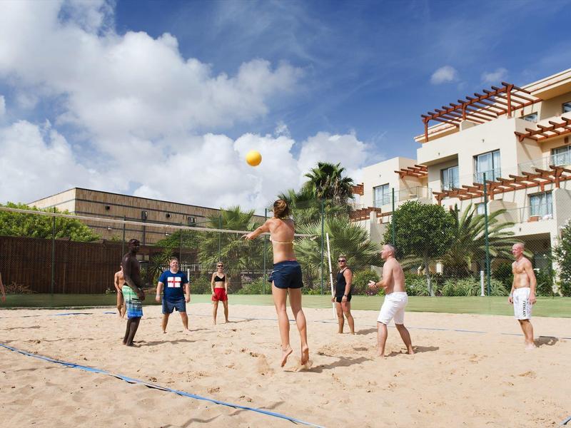 Mensen spelen beachvolleybal op een zandveld naast een hotel bij zonnig weer.
