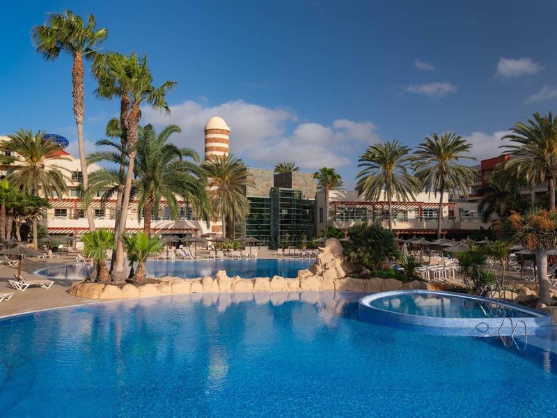 Groot hotelzwembad met parasols en palmbomen onder een blauwe lucht.