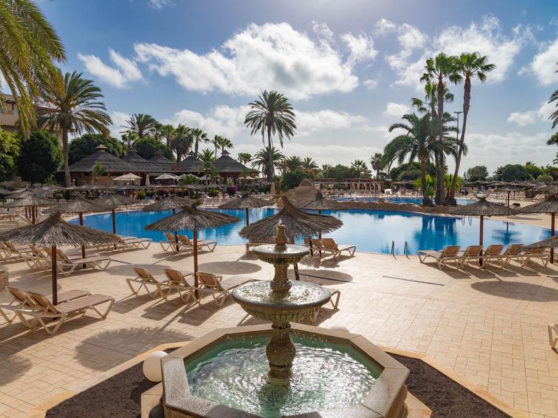 Groot buitenzwembad met palmbomen en parasols bij een hotelresort onder een blauwe lucht.