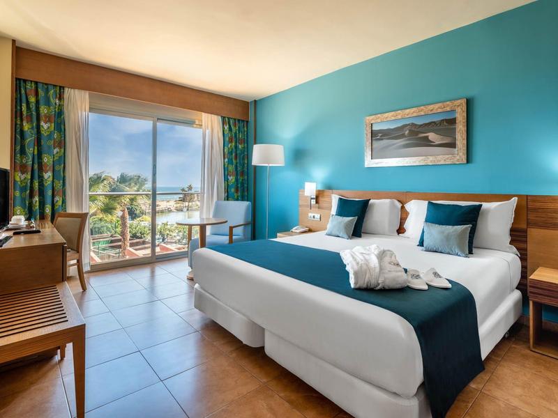 Lichte hotelkamer met tweepersoonsbed, balkon en zeezicht.