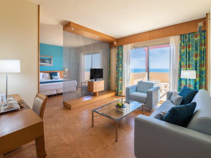 Lichte hotelkamer met bank, fauteuils, bureau en groot raam met uitzicht op zee.