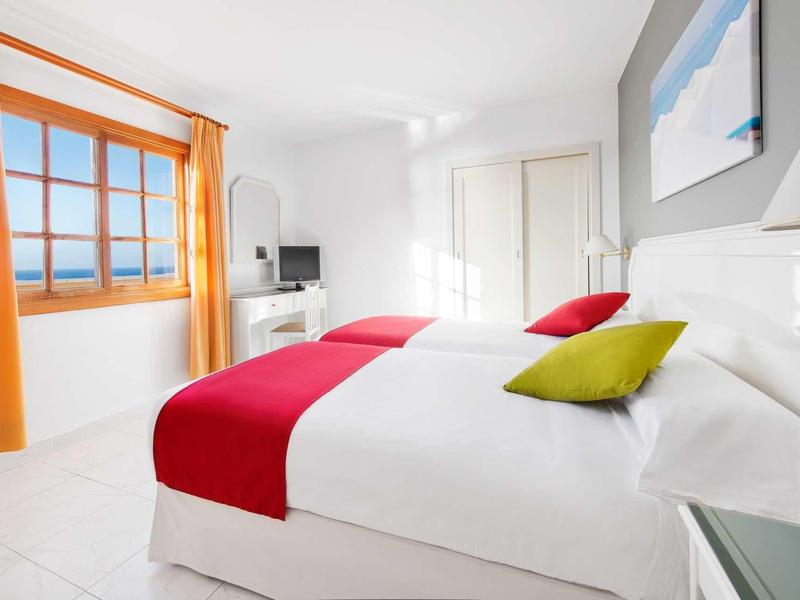 Světlý hotelový pokoj se dvěma postelemi, barevnými polštáři a výhledem na moře oknem.