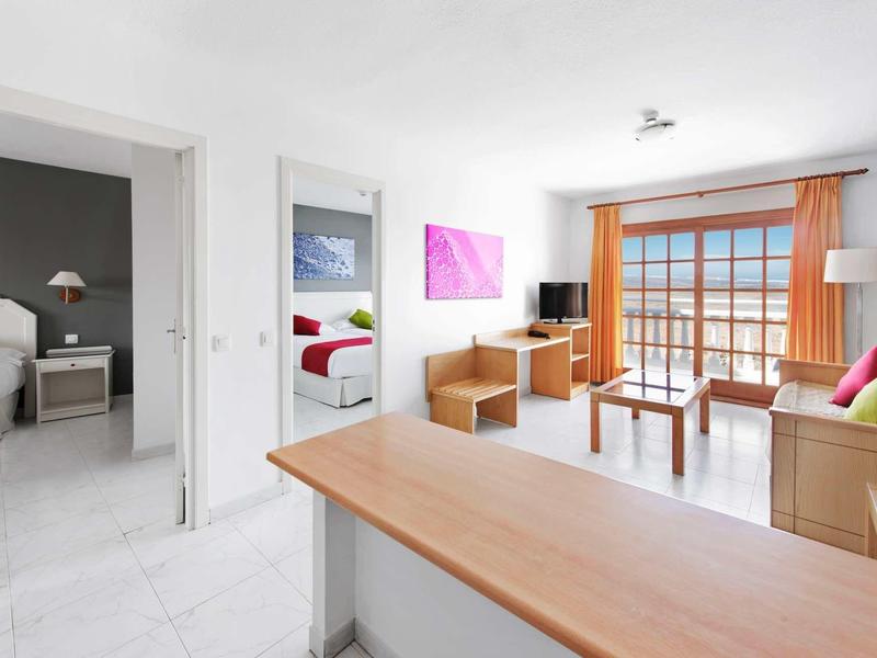 Světlá obývací část s dřevěným a bílým nábytkem, výhled do ložnice a na balkon.