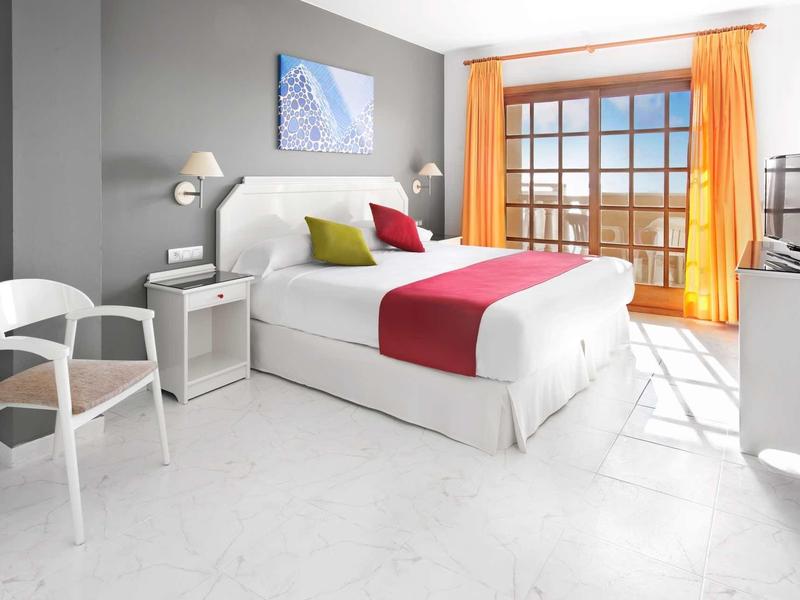 Světý hotelový pokoj s velkou postelí, dvěma židlemi, oranžovými závěsy a balkonem s výhledem na moře.