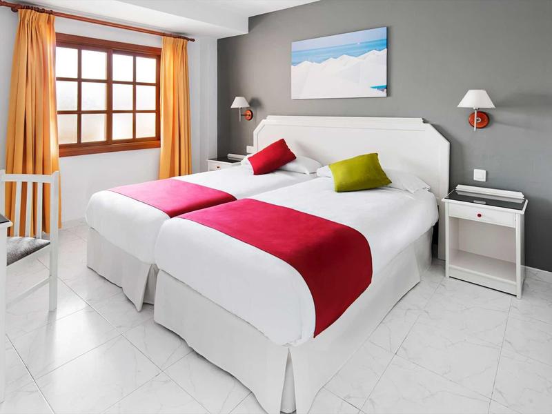 Moderní hotelový pokoj s manželskou postelí, bílým povlečením, barevnými polštáři a velkým oknem.