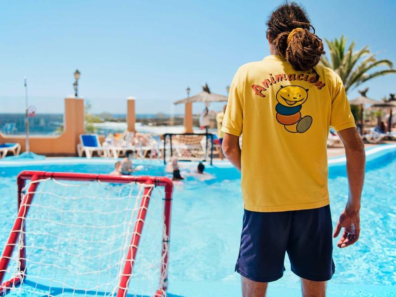 Muž v žlutém tričku a modrých šortkách stojí u bazénu a dívá se na slunečný bazén.