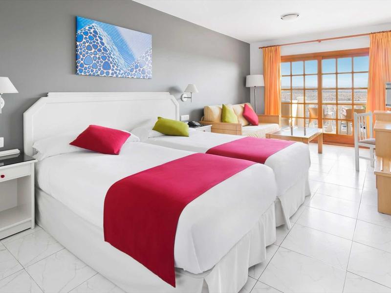 Světý hotelový pokoj se dvěma postelemi, barevnými akcenty, balkonem s výhledem na moře a moderním zařízením.