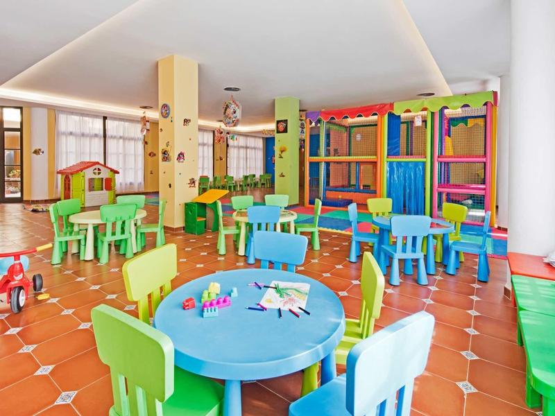 Barevná herna pro děti se stoly, židlemi a lezeckou konstrukcí v světlé místnosti.