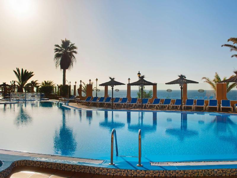 Velký hotelový bazén s lehátky, slunečníky a palmami u moře při západu slunce.