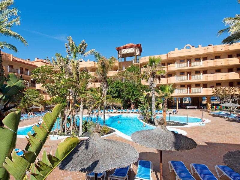 Venkovní bazén s lehátky a palmami před vícepodlažním hotelem za slunečného dne.