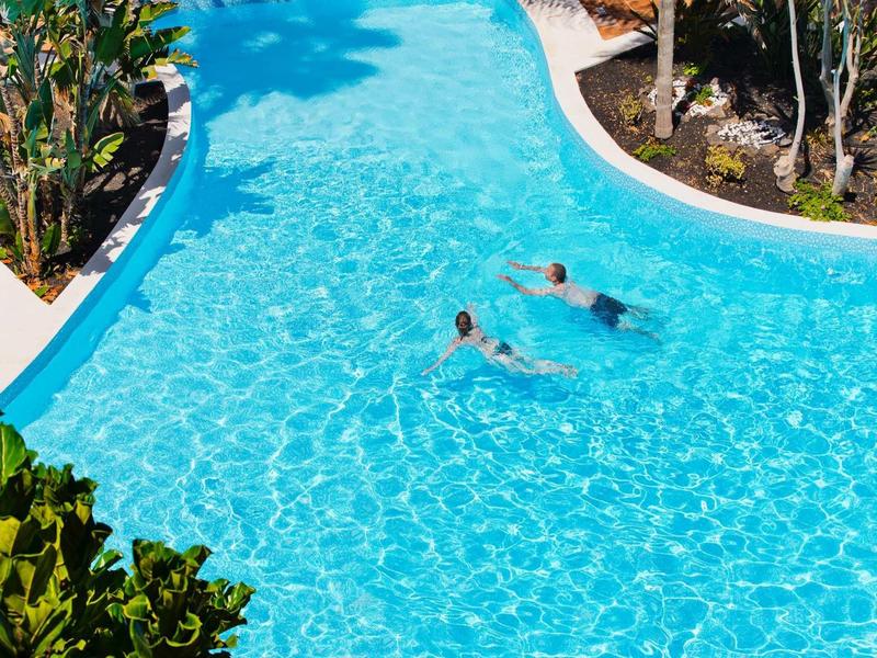 Dvě osoby plavou v čistém modrém bazénu s upravenou zahradou.