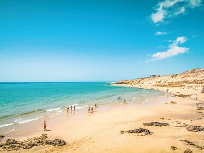 Strand mit goldenem Sand, blauem Meer und Menschen unter hellem Himmel mit vereinzelten Wolken.