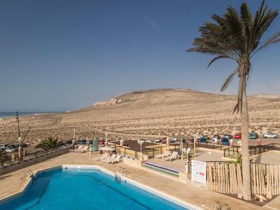 Blick auf einen blauen Pool mit Palmen, dahinter wüstenartige Landschaft und Gebäude unter klarem Himmel.