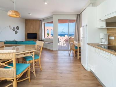 Helle offene Wohnzimmer-Küche mit Holzfußboden, blauem Sofa, Esstisch und Balkon mit Meerblick.