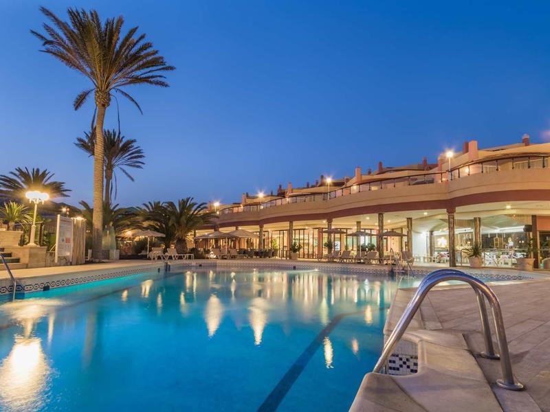 Area piscina dell'hotel illuminata con palme e posti a sedere al crepuscolo.