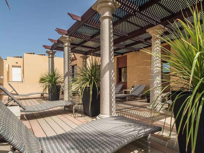 Terrazza soleggiata con lettini, colonne e grandi piante sotto un tetto di legno e vetro.