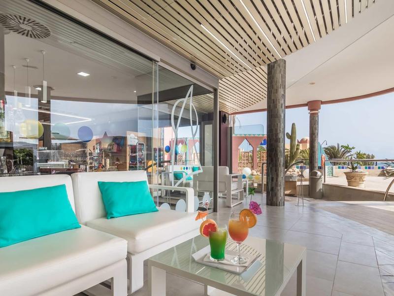 Area lounge moderna in hotel con divano bianco e bevande colorate su tavolo in vetro.