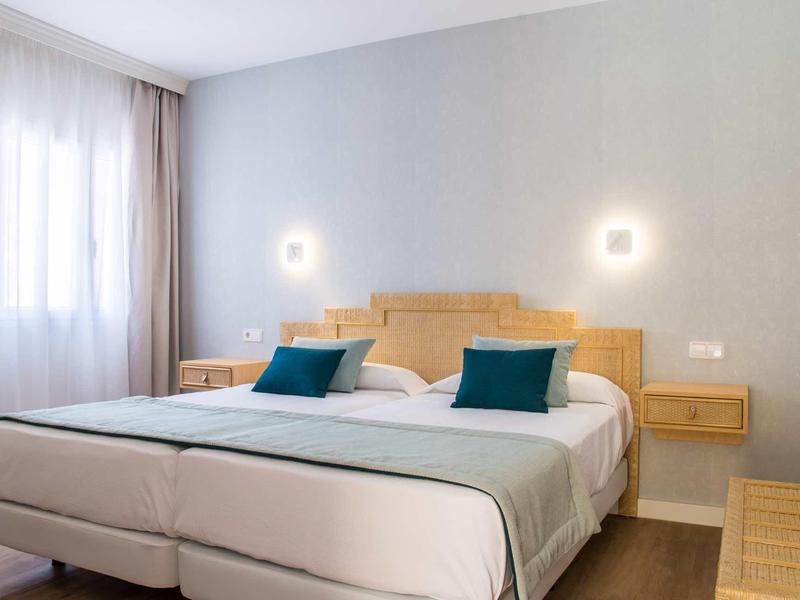 Camera d'hotel luminosa con due letti singoli, cuscini blu e comodini a muro.